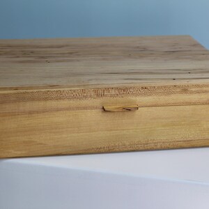 May include: A rectangular wooden box with a light tan grain and visible dark wood splines at the corners. The lid is flat and features a small, centred wooden tab handle on the front edge, showcasing natural wood textures and joinery details.