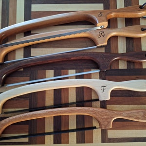 May include: Five wooden-handled saws with leather straps. The saws are arranged in a row, with the handles facing up. The handles are made of different types of wood, and each handle has a letter carved into it. The letters are S, A, L, F, and a cursive letter that is difficult to read.