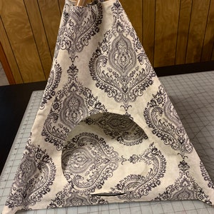 May include: A beige and brown patterned teepee tent for pets with a round opening. The teepee is made of fabric and has a wooden frame.