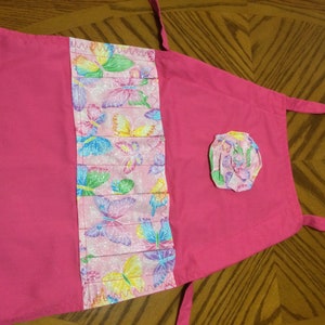 May include: Pink children's apron with a butterfly print and a pink fabric flower. The apron has a pocket with a pink and white butterfly print.