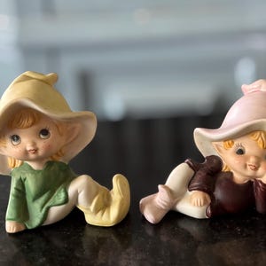 May include: Two ceramic figurines of elves, one with a yellow hat and green clothing, the other with a pink hat and brown clothing.