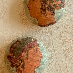 Peut inclure: Deux boutons ronds recouverts de tissu présentant des profils imprimés de personnages dans un style classique. Le bouton du haut montre une figure avec un couvre-chef à motifs, et le bouton du bas une figure avec un bandeau rouge, dans des tons terre.