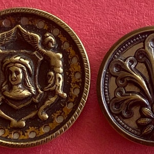 May include: Two antique brass buttons. One button features a woman's face surrounded by a shield and two figures. The other button has a stylised floral design.