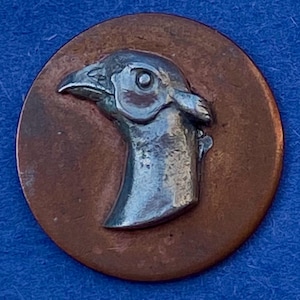 May include: A circular, copper-toned button featuring a raised, silver-coloured relief of a bird's head. The bird's head is detailed with a beak, eye, and feathers. The button has a rustic, vintage appearance.