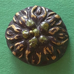 May include: A dark brown ceramic button with a raised floral design. The flower has five petals and is painted in shades of green and gold.