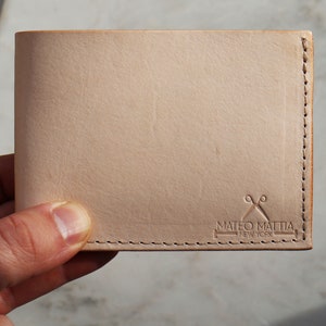 May include: A light brown leather wallet with a stitched edge and a logo that reads "Mateo Mattia New York" with a pair of scissors graphic.