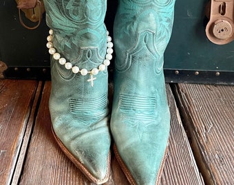 May include: A pair of teal leather cowboy boots featuring intricate tonal stitching and pointed toes. One boot is adorned with a white pearl anklet that includes a small gold-toned cross charm hanging from the center of the strand.