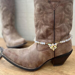 Könnte beinhalten: Braune Leder-Cowgirl-Stiefel mit einem weißen und goldenen Perlen-Knöchelband und einem großen Kristall-Verzierstück.