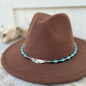 May include: A brown fedora hat with a decorative band featuring turquoise beads, silver beads, and a silver feather charm. The hat has a wide brim and a classic, stylish design.