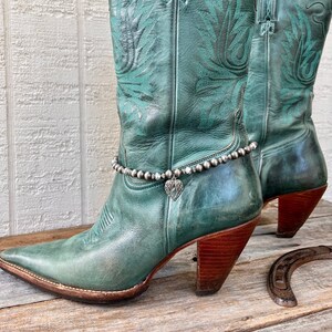 May include: Teal leather cowboy boots with a silver ankle bracelet featuring a heart charm with angel wings. The boots have a pointed toe and a high heel.