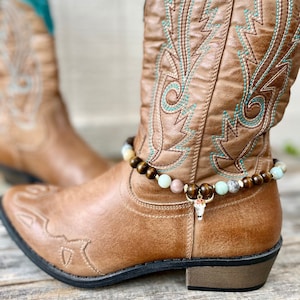May include: A pair of brown leather cowboy boots with turquoise accents and decorative stitching. A beaded ankle bracelet with a bull skull charm adorns one boot, showcasing a Western-inspired aesthetic.
