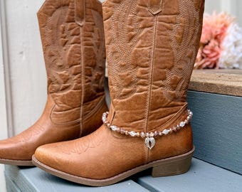 May include: A pair of tan leather cowboy boots featuring intricate stitched patterns on the shaft and toe. One boot is adorned with a beaded ankle bracelet made of clear and brown crystals, featuring a small silver-toned angel wing charm.