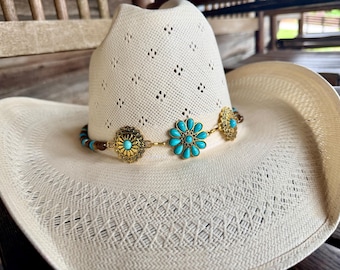 May include: A cream-colored straw cowboy hat featuring a decorative band made of gold-toned metal conchos and bright turquoise-colored stones. The hat has a woven texture with diamond-shaped ventilation holes throughout the crown and brim.