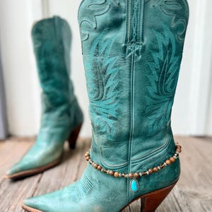 May include: A pair of teal leather cowboy boots featuring intricate tonal stitching on the shaft and a pointed toe. A beaded ankle bracelet with brown stone beads and a single teardrop-shaped turquoise pendant is wrapped around the front boot.
