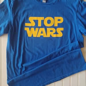 May include: A bright blue short-sleeved t-shirt featuring the text STOP WARS printed in a bold yellow font across the chest. The shirt has a classic crew neckline and a standard fit, with the graphic design centered on the front of the garment.