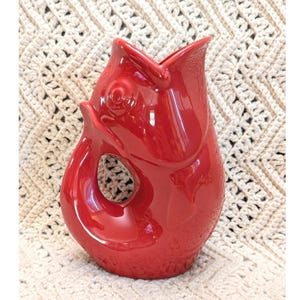 May include: A glossy, bright red ceramic pitcher shaped like a fish with its mouth open at the top. The handle is formed by the curved tail section of the fish, creating a circular opening in the center of the body. The surface is smooth and reflective.