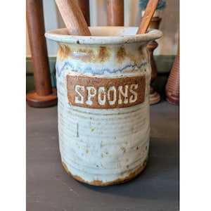 May include: A cylindrical ceramic utensil holder with a speckled off-white glaze and a brown rim. A rectangular brown plaque on the front features the word SPOONS in embossed white capital letters. Two wooden handles are visible inside.