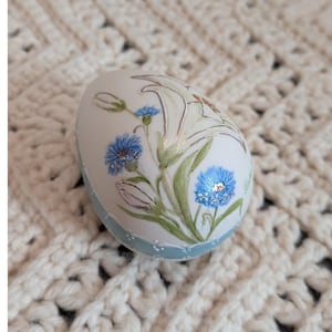 May include: A white, egg-shaped decorative item with a floral design. The egg features blue cornflowers and white lilies with green leaves. The base has a light blue band with white dots. The egg is resting on a cream-colored, crocheted surface.