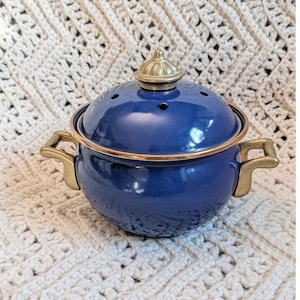 May include: A small, round, blue enamel pot with a lid and two handles. The pot has a gold-colored rim and handles. The lid has a gold-colored finial and several small holes. The pot is sitting on a white crocheted surface.