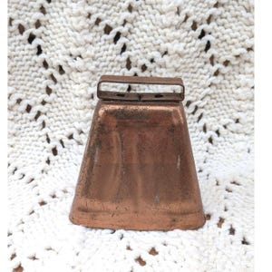 May include: A trapezoidal copper-colored metal bell with a weathered, oxidized surface texture. The bell features a flat, rectangular loop handle at the top and a wide, flared base, showcasing a distinct metallic sheen and signs of age.