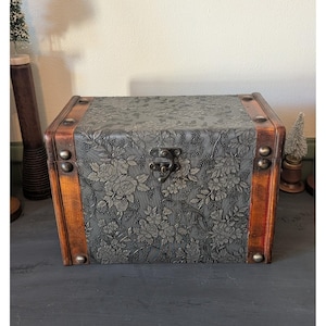 May include: A rectangular wooden storage chest featuring a dark gray embossed floral pattern on the front and top panels. The chest has smooth brown wooden edges, metal corner accents, and a central metal latch closure mechanism.