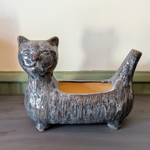 May include: A ceramic cat-shaped planter featuring a mottled blue, gray, and brown glaze with a glossy finish. The cat has a stylized face, a long body with a hollow center for plants, and a raised tail. The interior of the planter is tan.