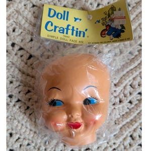 May include: A doll face in a clear plastic package. The doll face is peach-colored with blue eyes, black eyelashes, and red lips. The packaging reads "Doll Craftin' DIMPLE DOLL FACE 4IN".