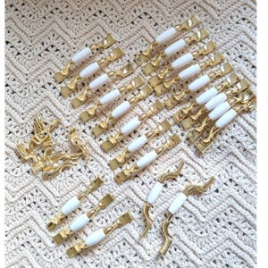 May include: A collection of gold-toned cabinet pulls and handles with white ceramic accents. The hardware features various designs, including bar pulls and decorative handles, arranged on a cream-colored crocheted surface.