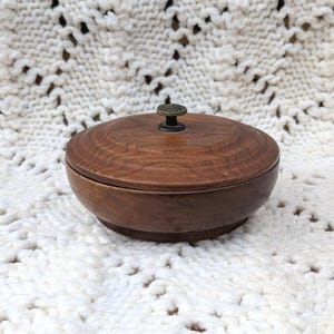 May include: A round, dark brown wooden bowl with a matching lid. The lid features a visible wood grain pattern and a small, centered, dark metallic knob handle. The bowl has a smooth, curved exterior and a flat base, presenting a simple, functional design.