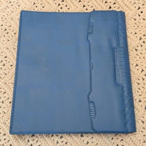 May include: A blue, textured binder with a snap closure. The binder has a ruler and other markings along the side. The binder is lying on a cream-colored, crocheted surface.