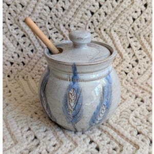 May include: A ceramic jar with a lid and a wooden spoon. The jar is light blue with darker blue wheat stalk designs and a matching lid. The jar is sitting on a cream-colored crocheted surface.