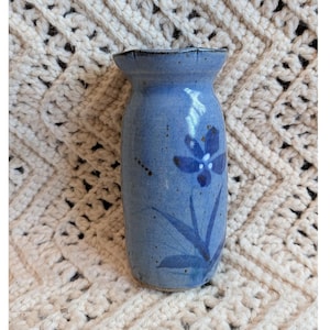 May include: A cylindrical ceramic vase with a speckled light blue glaze and a scalloped rim. The vase features a hand-painted dark blue floral design with leaves and a central flower, showcasing a smooth finish and a slightly flared top opening.
