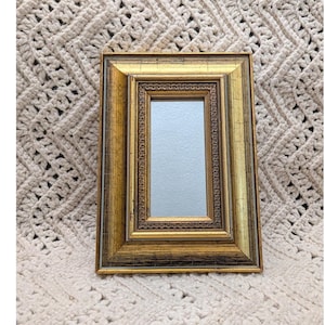 May include: A rectangular gold-colored picture frame with a mirror. The frame has a textured, distressed finish with dark brown accents. The mirror is rectangular and reflects a light-colored surface. The frame is resting on a beige crocheted blanket.