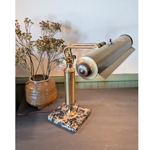 May include: A brass desk lamp featuring a cylindrical head with perforated ventilation holes, an adjustable arm, and a square base made of black and white speckled stone. The lamp has a metallic finish and a small black power switch.