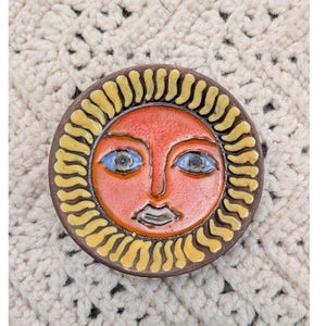 May include: A round, ceramic sun face with a brown border and yellow rays. The sun has a red face, blue eyes, and a small, dark mouth. The artwork is set against a cream-colored, textured background.