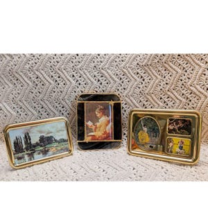 May include: Three decorative metal frames with gold-toned edges. The left frame displays a landscape painting. The center frame is black with a gold-toned border and shows a person reading. The right frame contains three separate small images.