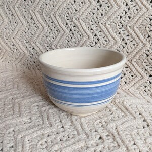May include: A ceramic bowl with a smooth, off-white base glaze featuring horizontal blue painted stripes around the exterior. The bowl has a rounded shape with a slightly flared rim and a clean, simple design that highlights the hand-painted finish.