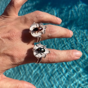 May include: Two silver rings, each featuring a clear, oval-shaped stone with a preserved insect inside. One insect is brown, the other is black. The rings are displayed on a hand against a blue water background.