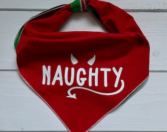 May include: Red pet bandana with white text reading "NAUGHTY" and a devil horn and tail graphic. The bandana has a green and black trim and is tied at the top. The background is a white wooden surface.