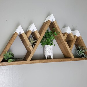 May include: A wooden mountain-shaped shelf with white painted peaks. The shelf has five compartments and is decorated with small potted plants.