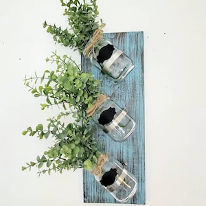 May include: Three glass jars with black chalkboard labels are attached to a rustic blue wooden plank. Each jar is filled with artificial greenery.