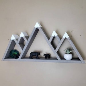 May include: Gray wooden mountain-shaped shelf with white peaks. The shelf has three tiers and is decorated with small toy bears and a potted succulent.