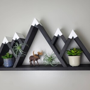 May include: Black wooden mountain-shaped shelf with white painted peaks. The shelf has three tiers and is decorated with potted plants and a small moose figurine.