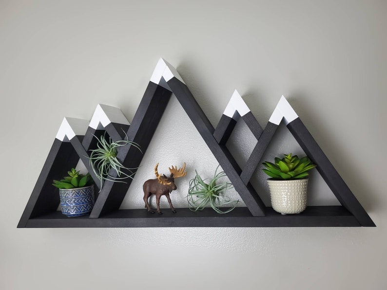 May include: Black wooden mountain-shaped shelf with white painted peaks. The shelf has three tiers and is decorated with potted plants and a small moose figurine.