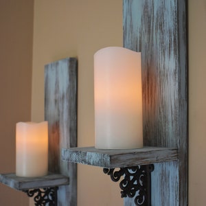 Peut inclure: Deux appliques murales rustiques avec des panneaux arri&egrave;re en bois bleu-gris vieilli et de petites &eacute;tag&egrave;res. Chaque &eacute;tag&egrave;re contient une grande bougie pilier blanche et brillante. Des supports en m&eacute;tal noir orn&eacute; soutiennent les &eacute;tag&egrave;res.