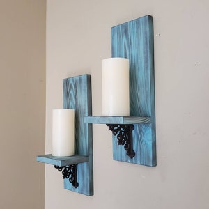 Peut inclure: Deux bougeoirs muraux avec une finition bleu vieilli. Chaque support comporte une petite &eacute;tag&egrave;re soutenue par des supports m&eacute;talliques noirs orn&eacute;s. Des bougies piliers blanches reposent sur les &eacute;tag&egrave;res, cr&eacute;ant un affichage d&eacute;coratif rustique.
