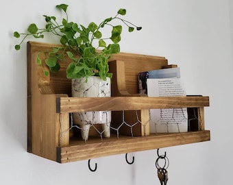 Può includere: Mensola da parete in legno rustico con un inserto in rete metallica, tre ganci e una piccola pianta in vaso. La mensola ha un cartello con la scritta "Go Homestead for the Holidays".