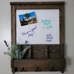 May include: A rectangular wall-mounted whiteboard framed in dark stained wood with a storage shelf, three black metal hooks, and a small bouquet of lavender. The board displays handwritten text: Have a great day!, Dentist Appt. @ 3PM, -Milk, -Bread, -Eggs.