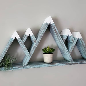 May include: A teal blue wooden mountain shelf with white peaks. The shelf has three triangular shelves and two small potted plants.