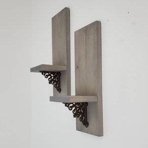 Peut inclure: Deux &eacute;tag&egrave;res flottantes en bois gris avec des supports d&eacute;coratifs en m&eacute;tal fonc&eacute;. Les &eacute;tag&egrave;res sont mont&eacute;es sur un mur blanc, mettant en valeur un design rustique. Chaque &eacute;tag&egrave;re a une forme rectangulaire et est soutenue par des supports orn&eacute;s.